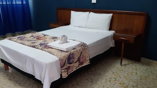 A neatly made bed with a floral bedspread, a white pillow, and a small table with a rolled-up towel and a bottle. - Hotel Narváez Classic (Tuxtla Gutierrez)