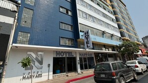 Un hotel de varios pisos con fachada azul, entrada visible y zona de estacionamiento con automóviles.