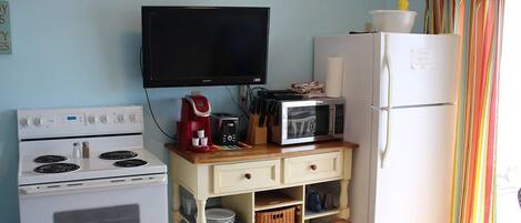 Una cocina compacta con estufa, microondas y refrigerador, un televisor sobre la estufa, y una ventana con cortinas.