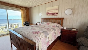 Une chambre avec un grand lit, une tête de lit en bois, une table de chevet, une chaise, un ventilateur et une vue sur la mer par la fenêtre.