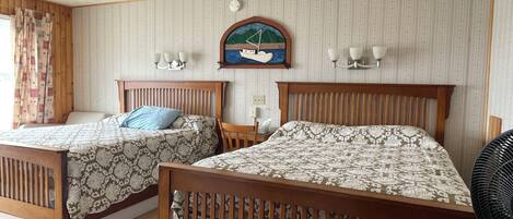 A room with two wooden beds, a wall-mounted painting of a sailboat, a small table, and a wall-mounted fan.