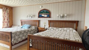 Une pièce avec deux lits en bois, un tableau représentant un voilier accroché au mur, une petite table et un ventilateur fixé au mur.