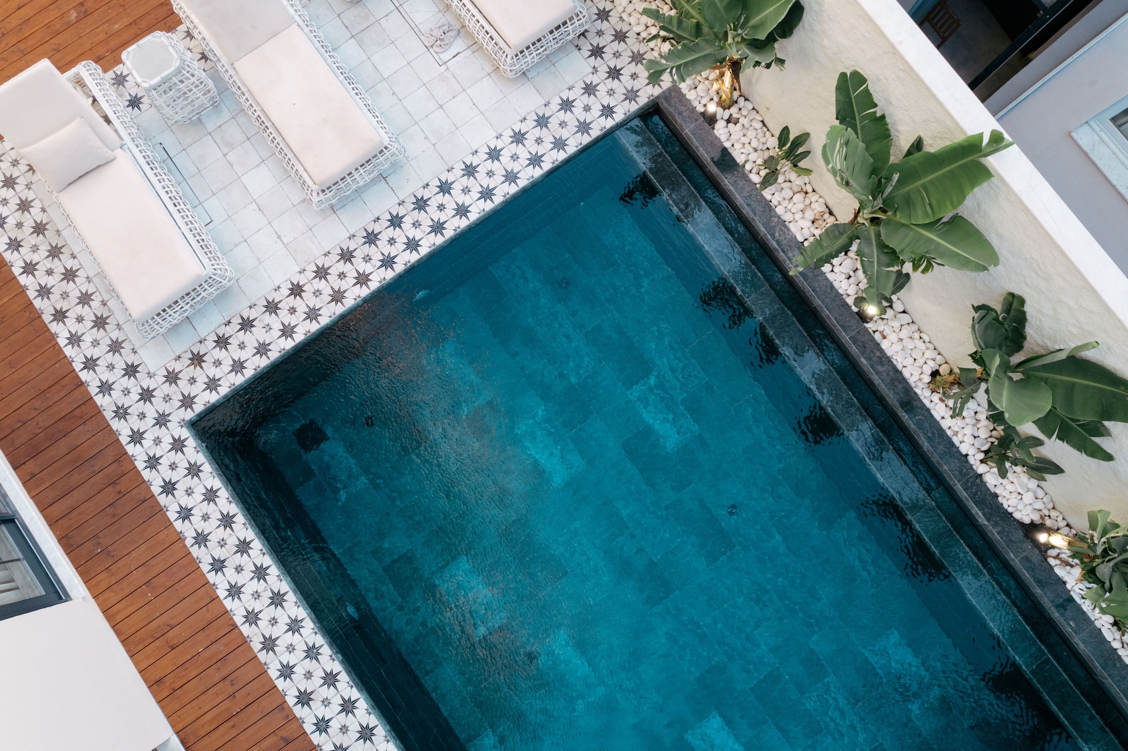 A rectangular pool with a tiled border, surrounded by white lounge chairs and a small garden area with plants.