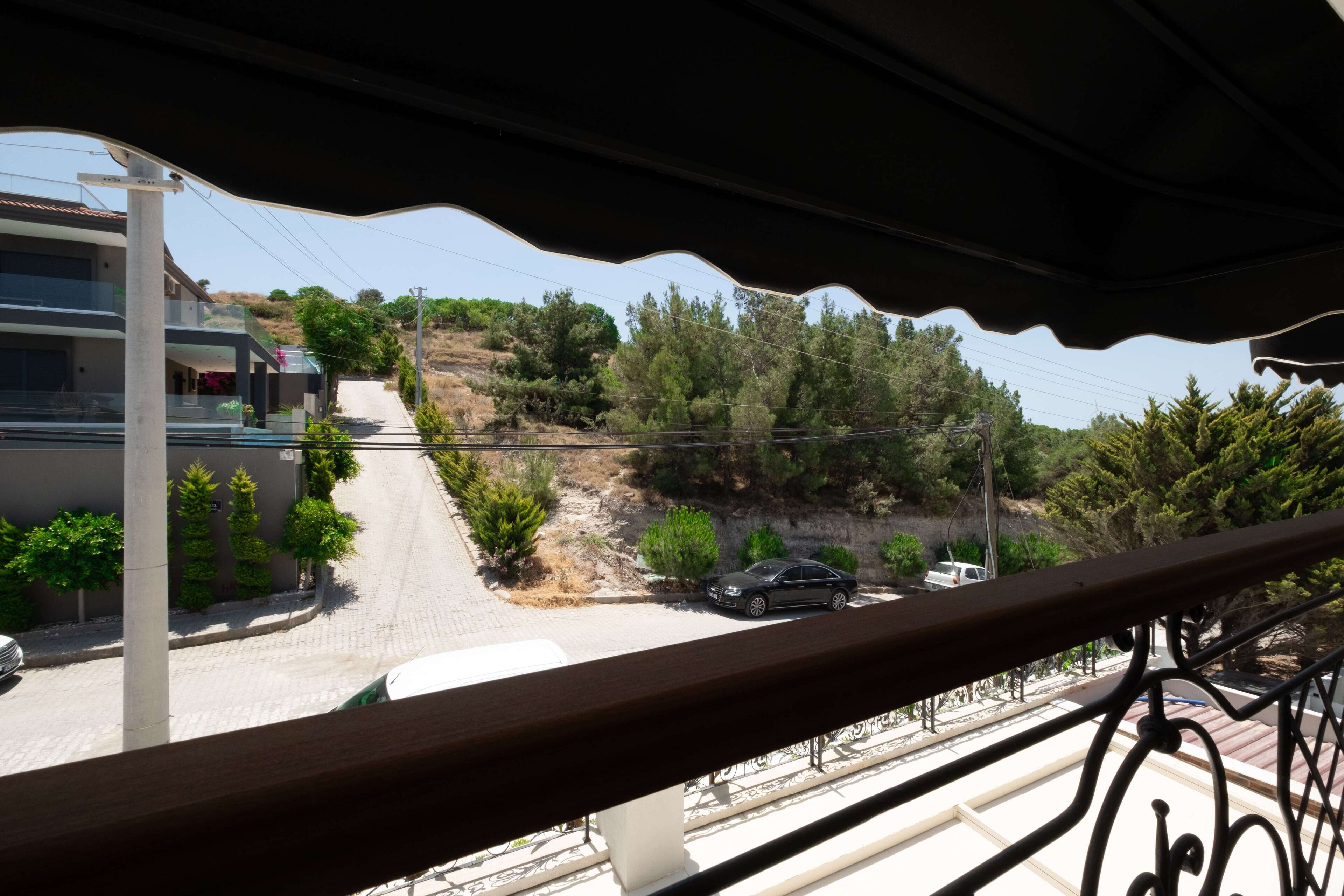 A balcony with a view of a residential street, a parked car, and a hillside with trees.
