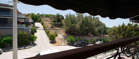 A balcony with a view of a residential street, a parked car, and a hillside with trees.