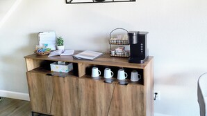 A wooden cabinet with a coffee maker, mugs, a plant, and a small table with a book and a basket.