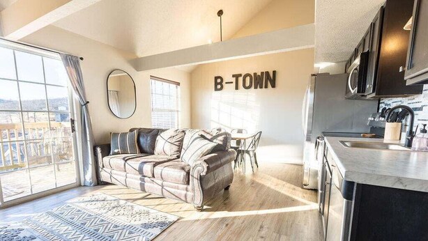 A modern living room with a sofa, a coffee table, and a rug. There is a kitchen area with a refrigerator and a microwave. The room has a large window and a wall with the text "B-TOWN".