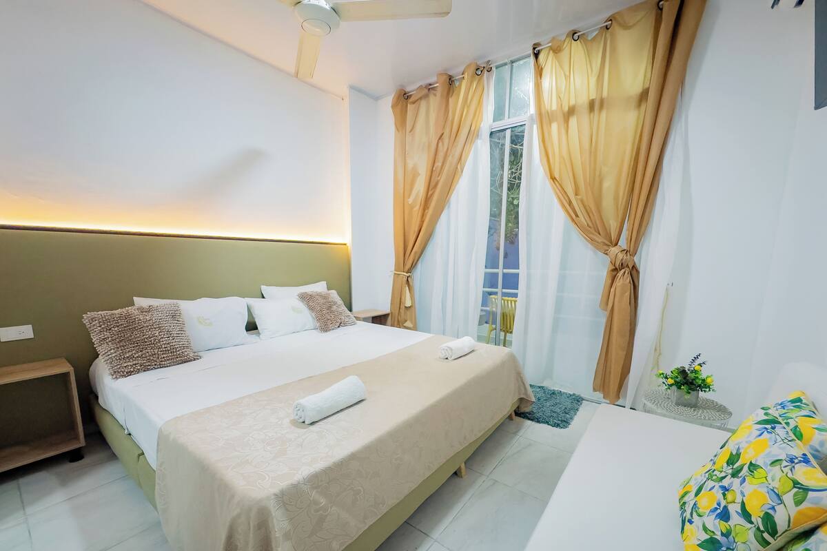 A hotel room with a bed, a nightstand, a window with curtains, and a small table with a plant.