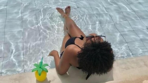 A person lying on a poolside with clear water, a small yellow plant, and tiled flooring.