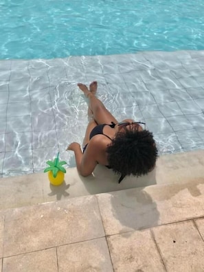 Una persona estesa al costat d'una piscina amb aigua cristal·lina, una petita planta groga i paviment de rajoles.