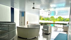 A modern hotel lobby with a large, curved white bathtub, a seating area with white armchairs, and a view of lush greenery outside.