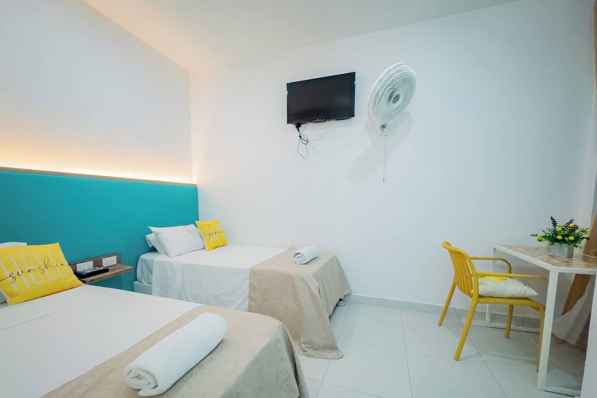 A hotel room with two beds, a desk, a chair, a TV, and a wall-mounted fan.
