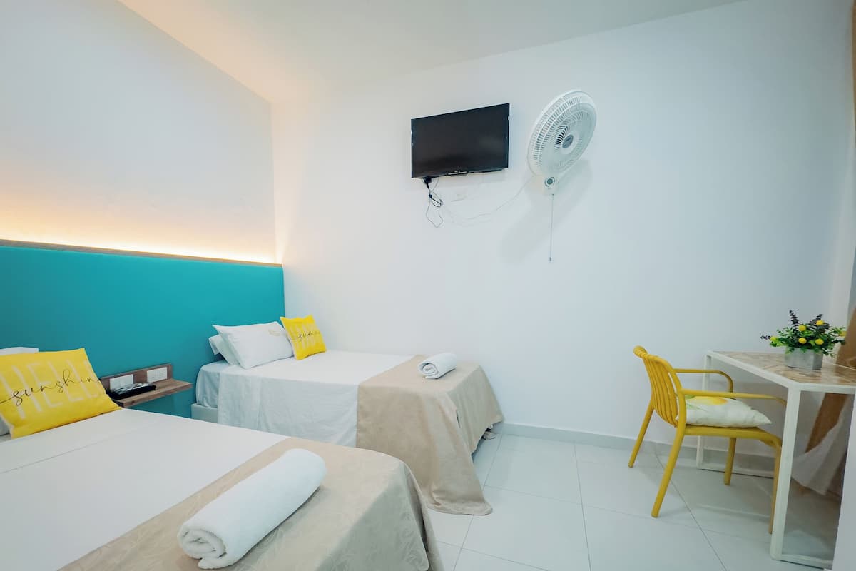 A hotel room with two beds, a desk, a chair, a TV, and a wall-mounted fan.