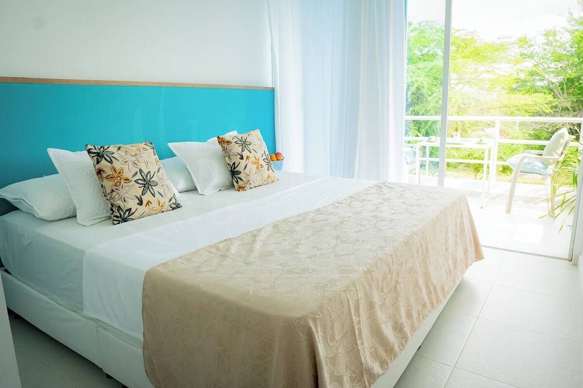 A hotel room with a bed, a balcony with a chair, and a view of greenery.