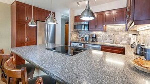 A modern kitchen with granite countertops, stainless steel appliances, wooden cabinets, and pendant lighting. - Oceanfront, King Bed, Split AC, WiFi, Hot Tub Pool (Wailuku)