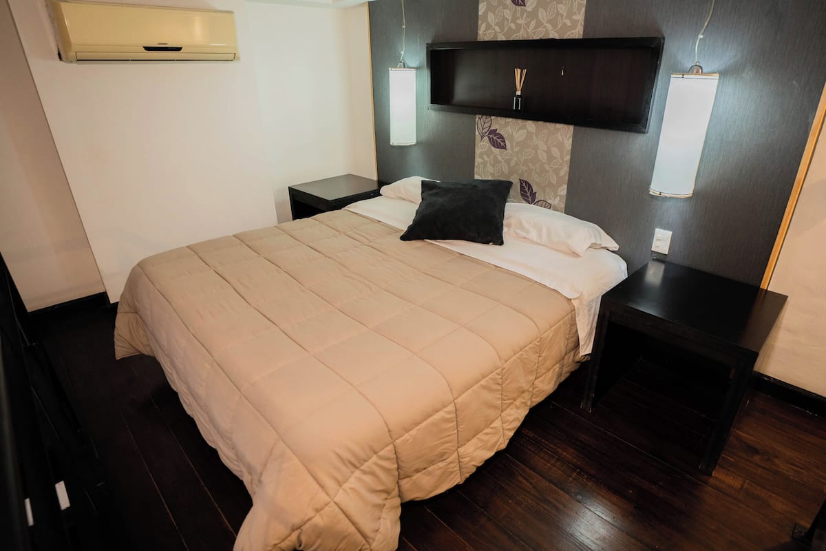 A neatly made bed with a beige duvet, a black pillow, and a headboard with a shelf. There are two bedside tables, one with a lamp and the other with a small item on it. The room has dark wooden flooring and a wall-mounted air conditioner.