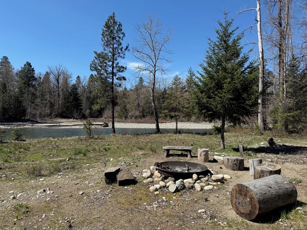 A campsite with a fire pit, logs, rocks, and a river in the background.