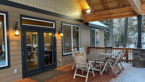A covered deck with a wooden table and chairs, a stone fireplace, and a view of snow-covered trees.
