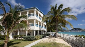 Un hotel frente a la playa con fachada blanca, varios balcones y un camino de arena que conduce a la orilla.