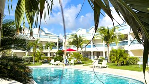 Un área de piscina con sillones de descanso y una piscina azul cristalina, rodeada de palmeras y un edificio con balcones.
