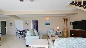 Una sala de estar con sofá, sillón, mesa de comedor y sillas. Hay un televisor y un jarrón sobre una mesita.