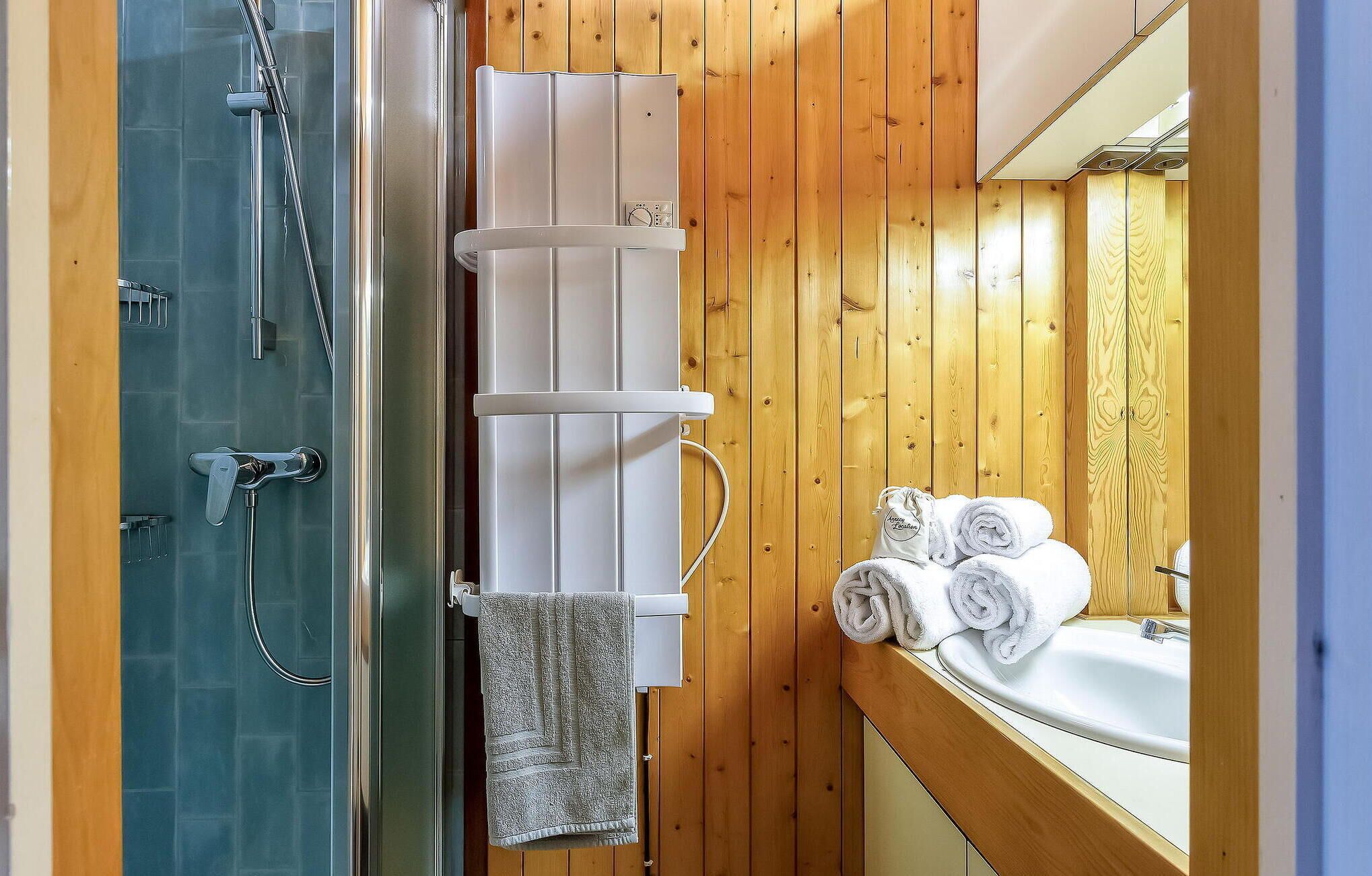 A bathroom with a glass shower enclosure, a white towel rack, and a sink with rolled towels.