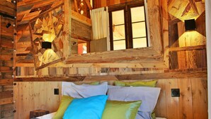Interior de una cabaña de madera con una cama, almohadas y dos lámparas fijadas a la pared.