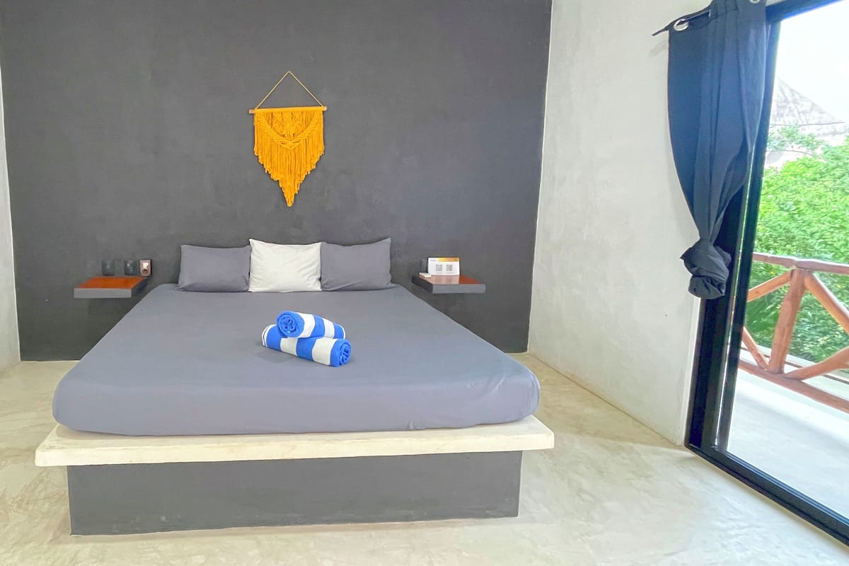 Un lit avec un matelas gris, des oreillers et une serviette bleue et blanche. Il y a une petite table avec un livre et une lampe de chaque côté du lit. Une décoration jaune est suspendue au mur, au-dessus du lit.