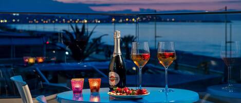 A table with a bottle of Martini wine, two glasses of wine, a plate of strawberries, and two lit candles on a balcony overlooking a body of water and mountains at dusk.