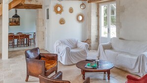 A spacious living room with a high ceiling, exposed wooden beams, a dining area with a table and chairs, and a large window providing natural light.