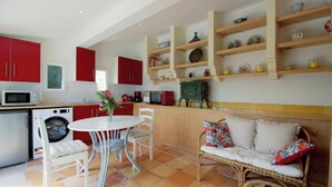 Une cuisine compacte avec des meubles rouges, une table à manger avec des chaises, un lave-linge et un canapé en osier avec des coussins.