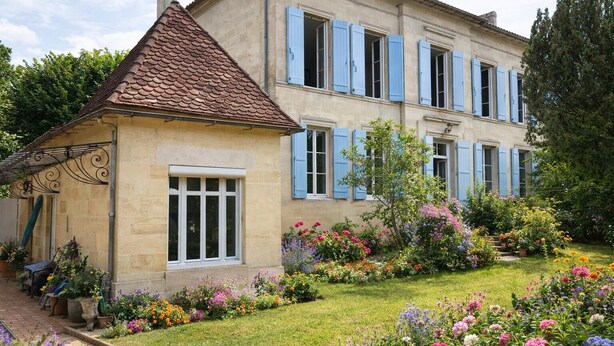 Une maison à deux étages, avec un toit en tuiles, une petite dépendance attenante et un jardin où poussent différentes sortes de fleurs.