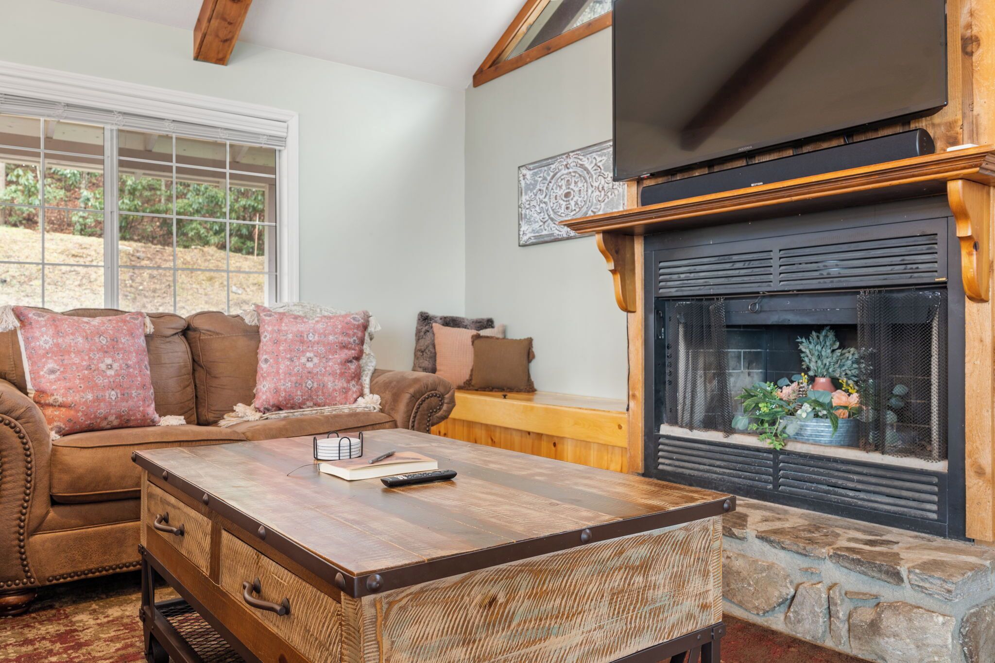 A cozy living room with a fireplace, a sofa with patterned cushions, and a coffee table with a book and remote control.