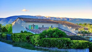 A two-story house with a tiled roof and green shutters, surrounded by lush greenery and mountains in the background. - Les Murmures Du Ventoux (Mormoiron)