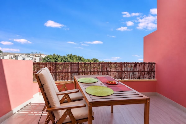 Un balcón con una mesa de madera y dos sillas, un muro rojo, y vistas a edificios y árboles.