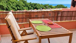 A wooden table with green placemats and a plate of food, a cushioned chair, and a view of the ocean through a bamboo fence.
