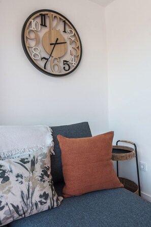 Una habitación con un reloj de pared redondo, un sofá gris y una pequeña mesita de noche con una cesta.