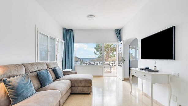 A modern living room with a beige sofa, a flat-screen TV mounted on the wall, and a view of the outdoors through large windows.