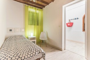 A bedroom with a bed, a chair, a window with curtains, a door, and a wall-mounted coat rack with bags. - Home2Book Oceanfront Bliss in Taliarte (Taliarte)