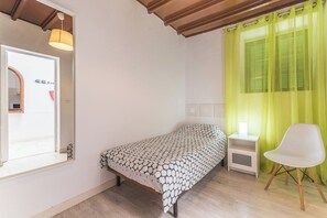 A single bed with a patterned bedspread, a bedside table with a lamp, a chair, and a window with green curtains.