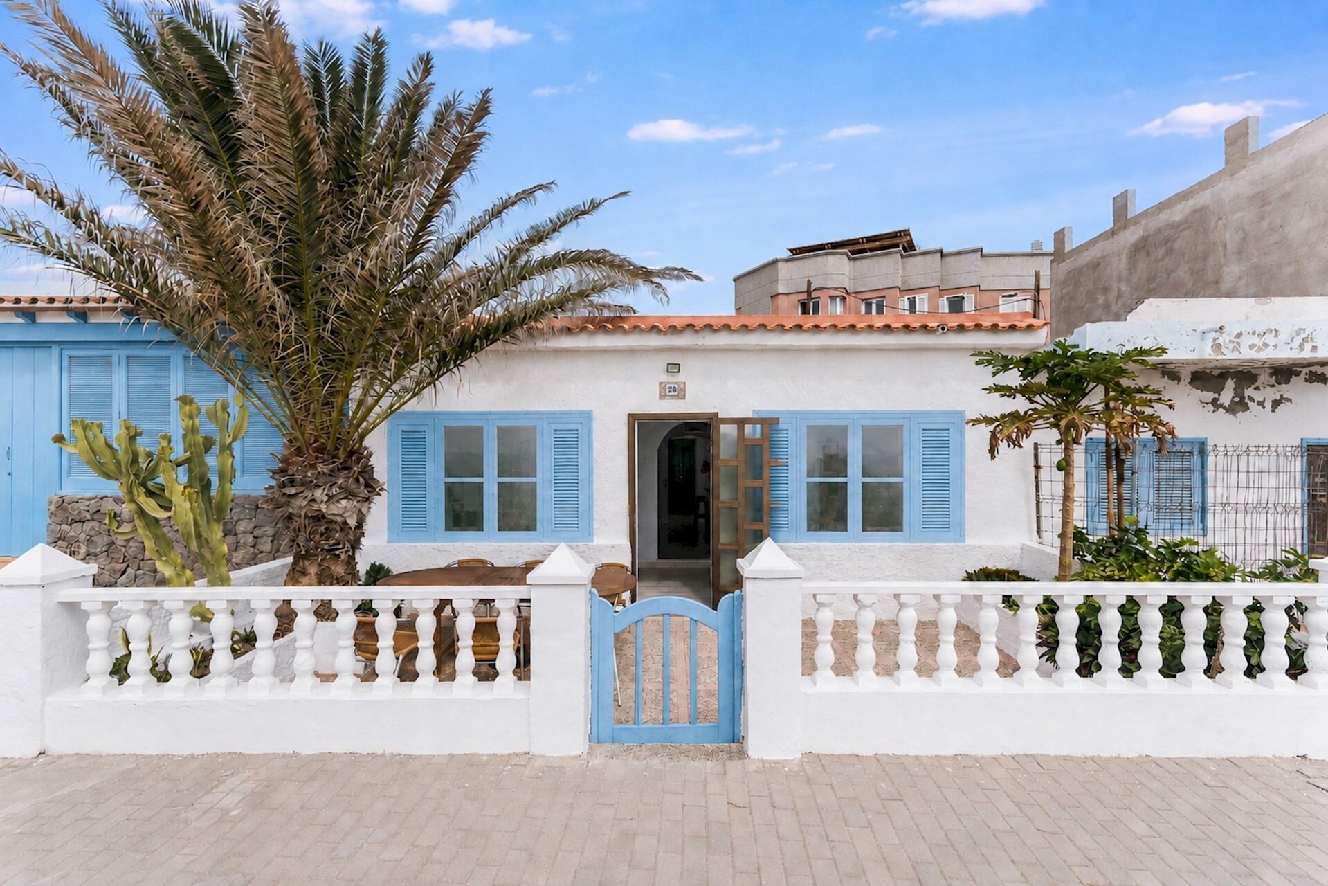 Un edificio blanco con contraventanas azules, una reja y una palmera.