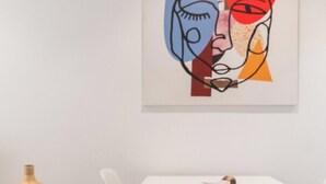 Un coin repas minimaliste avec une table et des chaises blanches, un vase en bois posé sur un piédestal et une peinture abstraite moderne accrochée au mur.