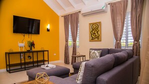 A living room with a large gray sofa, a wooden coffee table, a TV mounted on the wall, and a small side table with a plant. - Sunshine Paradise Villa (St. Ann's Bay)