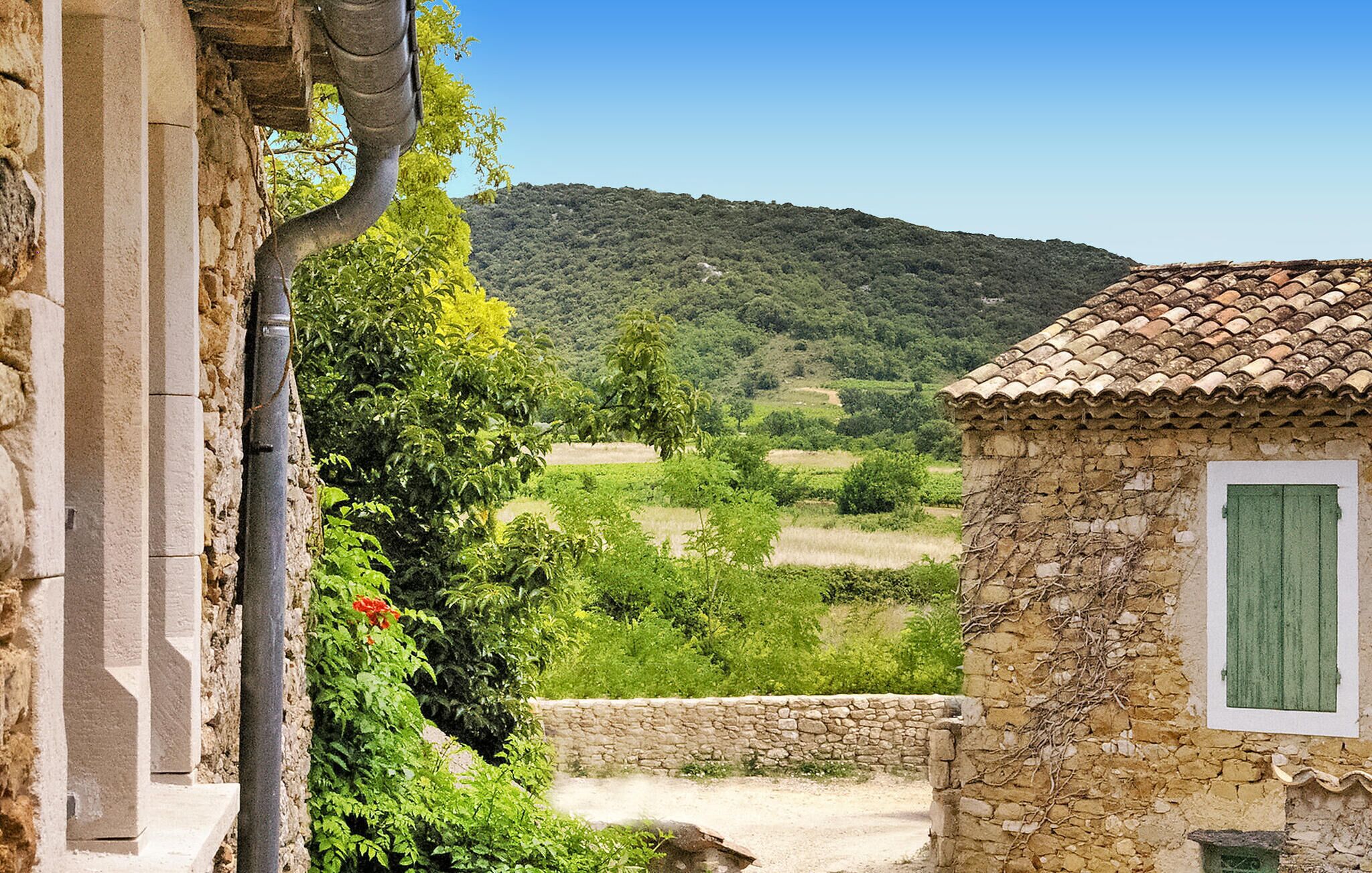 A rustic stone building with a tiled roof, a stone wall, and a view of a green valley and hills.