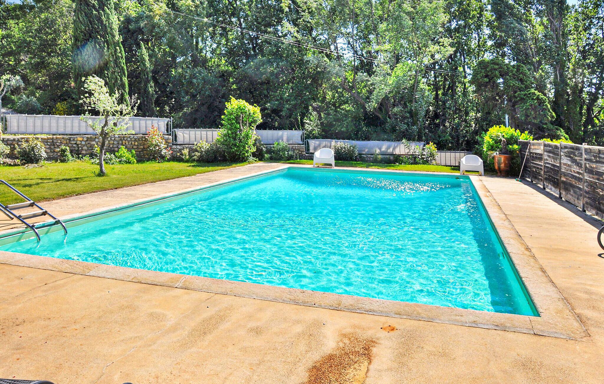 A rectangular swimming pool with a ladder on one side, surrounded by a stone wall and a grassy area with trees and a wooden fence.