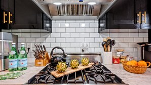 Une cuisine équipée d’une cuisinière à gaz, une planche à découper en bois ornée d’artichauts, un panier d’oranges et des bouteilles d’eau Pellegrino.