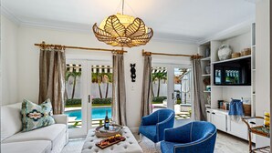 Un salon avec un canapé blanc, des fauteuils bleus, une table basse en bois sur laquelle on a disposé des fruits, et un téléviseur posé sur un meuble blanc.