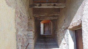 Un étroit couloir en pierre, avec un sol carrelé, une porte en bois sur la droite et une fenêtre au fond.
