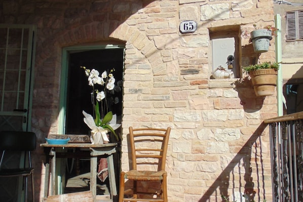 A stone building with a number '65' sign, a wooden chair, a table with a blue bowl, and a potted plant.