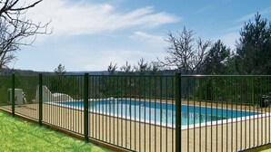 A rectangular swimming pool surrounded by a metal fence, with a grassy area in the foreground and trees in the background.